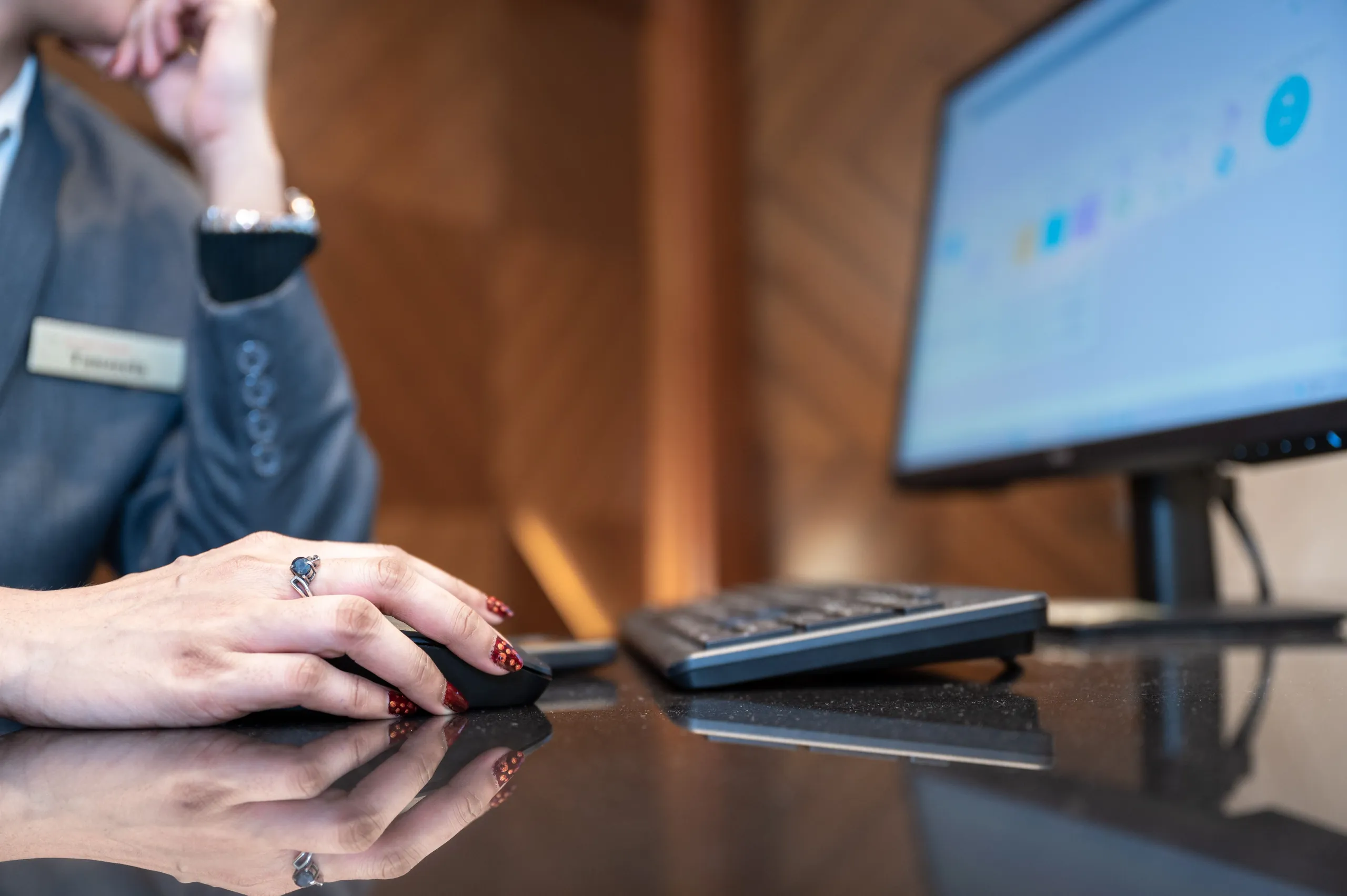 Close-up of a receptionist's hand on a mouse at a polished desk, with a keyboard and a monitor showing a scheduling screen beside it.