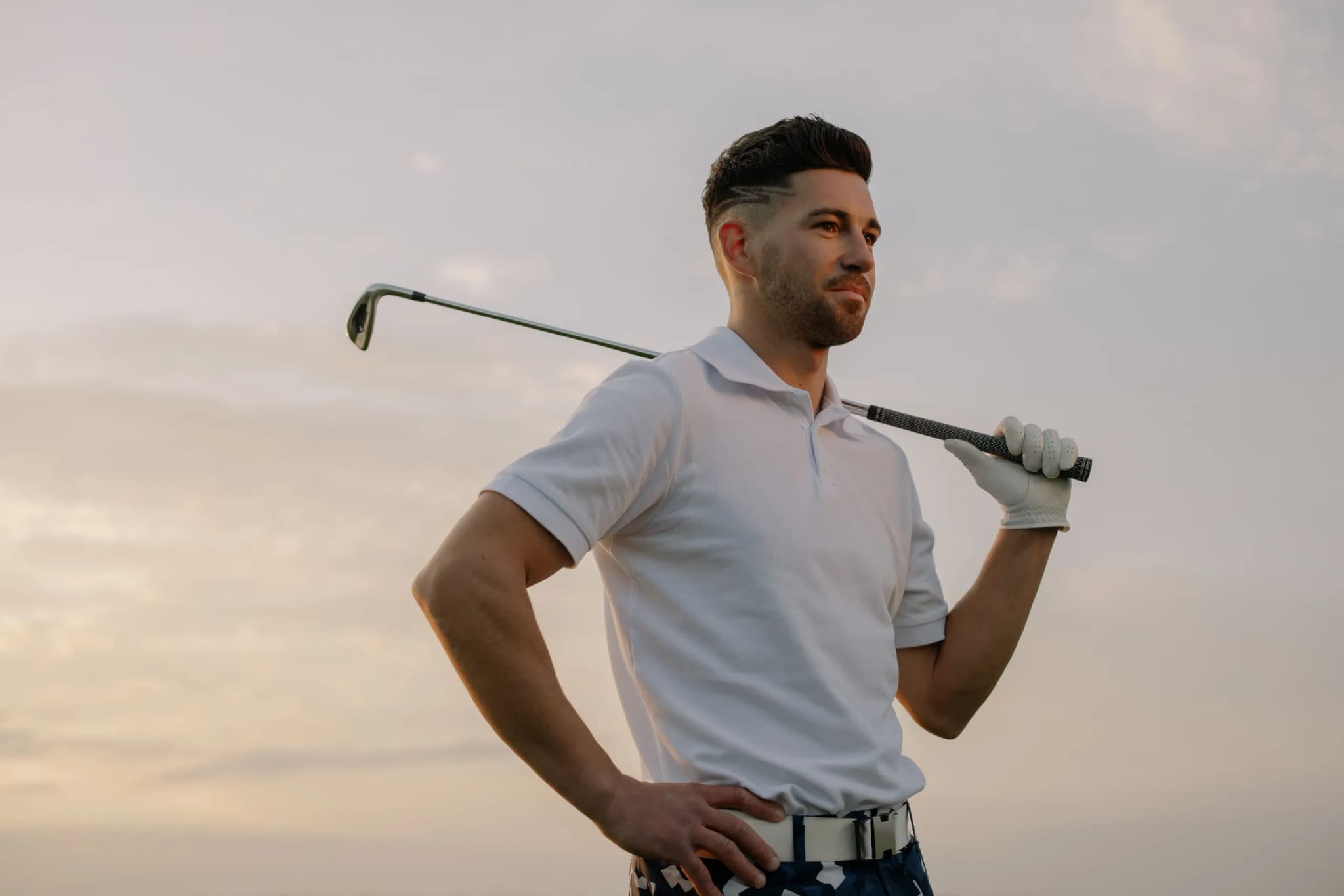 A golfer in a white polo standing against a pale evening sky, an iron resting across one shoulder.