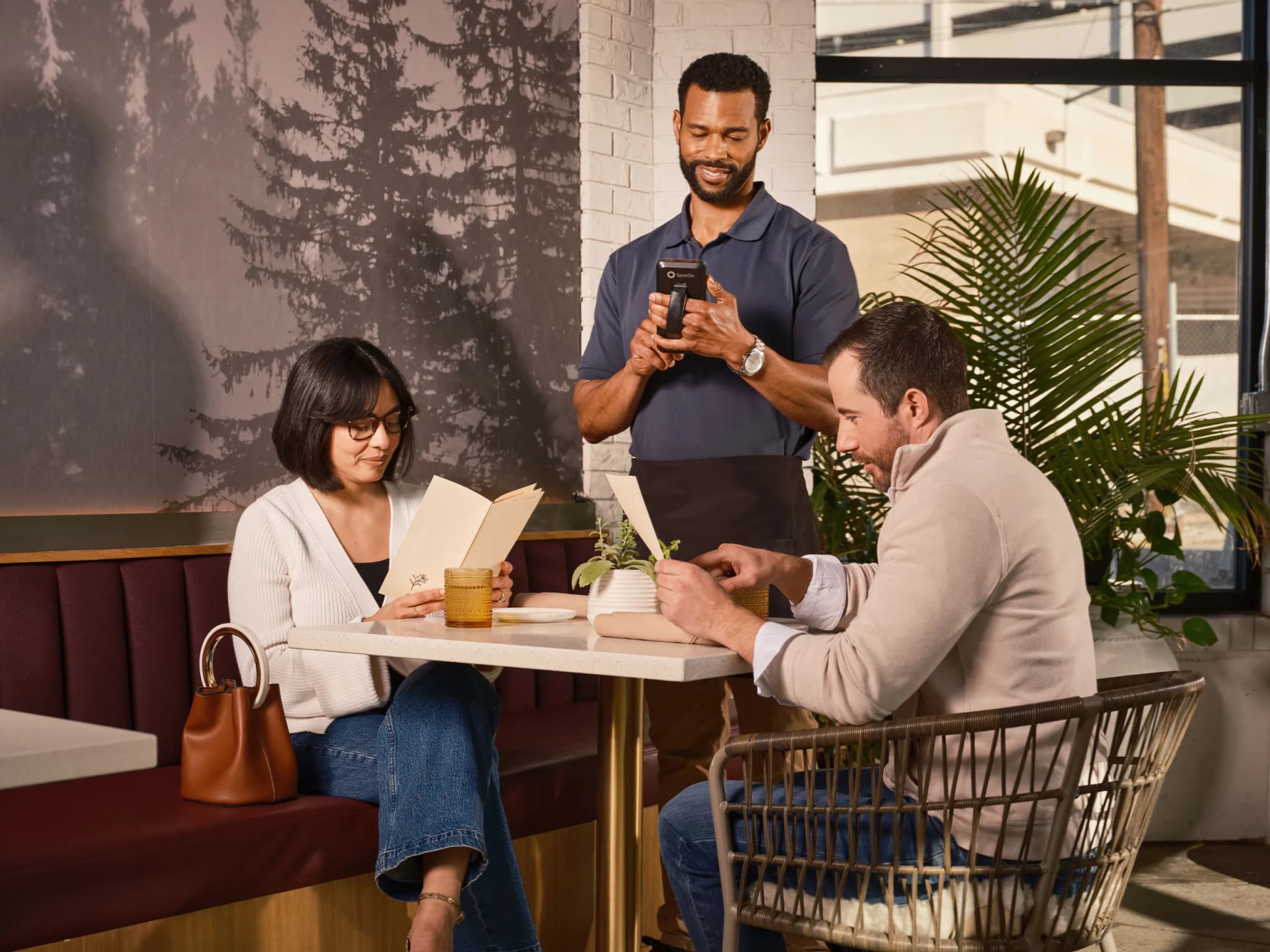 Three people around a cafe table, one taking an order on a handheld card terminal while the other two sit with a book and drinks.