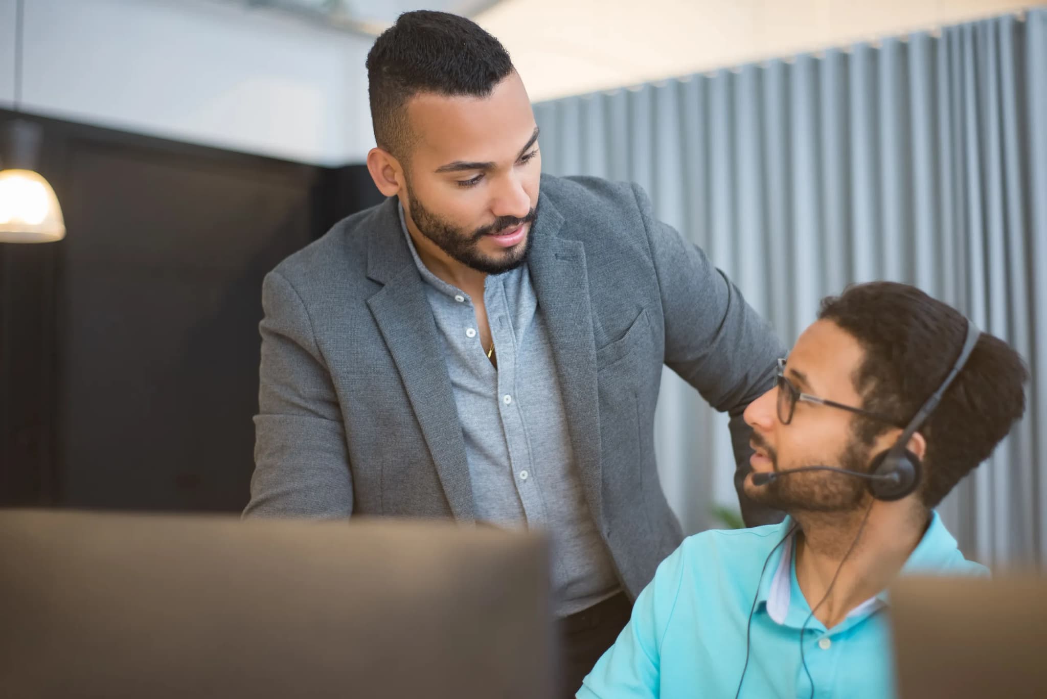 Two colleagues in an open-plan office, one leaning in to talk with a support agent seated at a monitor wearing a headset.