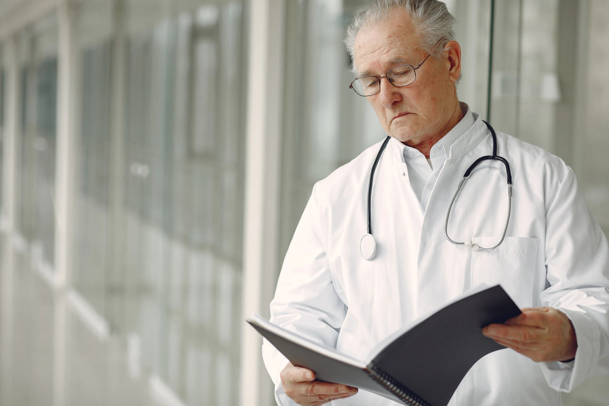 A doctor in a white coat and stethoscope reading a patient file in a bright hospital corridor.